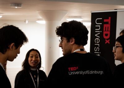 Students chatting in a huddle whilst wearing black t-shirts with TEDxUniversityofEdinburgh on them.