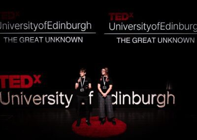 A male (me) and female, both in their twenties, stand on a professional stage with signs of "TEDxUniversityofEdinburgh" behind them.