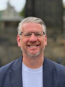 Man with grey hair, glasses, and a beard smiling.